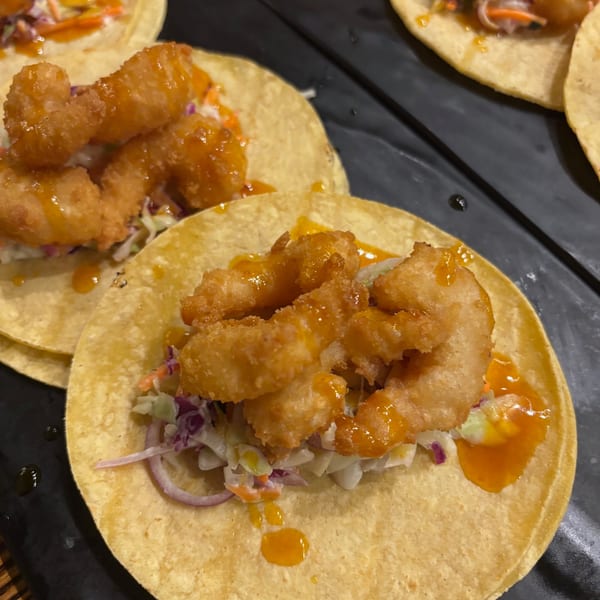 Close-up of Uncommon Grill's crispy shrimp tacos with slaw and orange sauce on a dark surface.