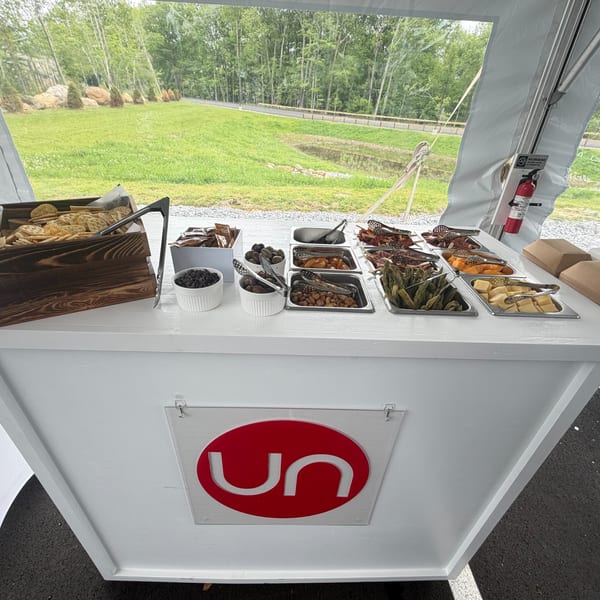 A white buffet table with various snacks and appetizers under a tent, with a green landscape outside.