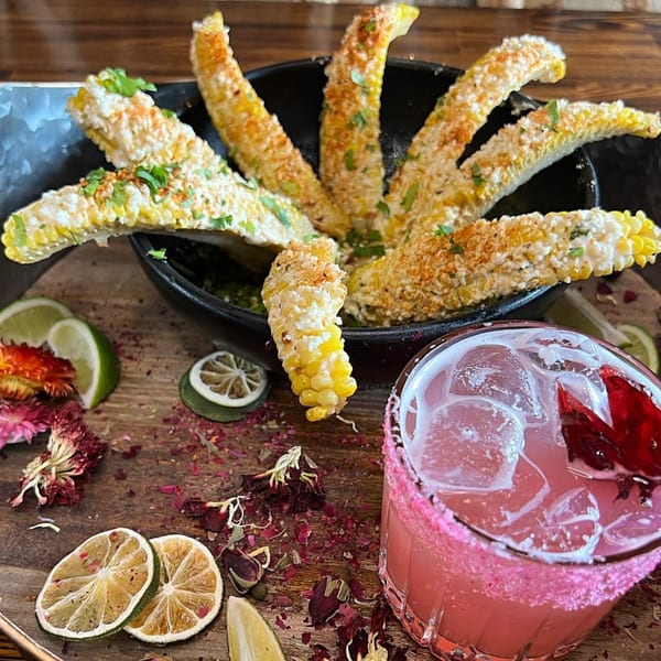 Elote corn ribs and a pink margarita with a sugared rim on a wooden tray with dried flowers.