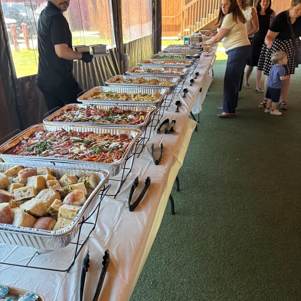 A long buffet table filled with hot food in aluminum trays, with people serving themselves.