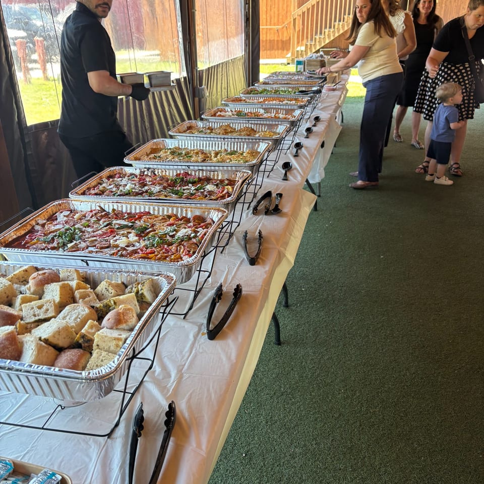 A long buffet table filled with hot food in aluminum trays, with people serving themselves.
