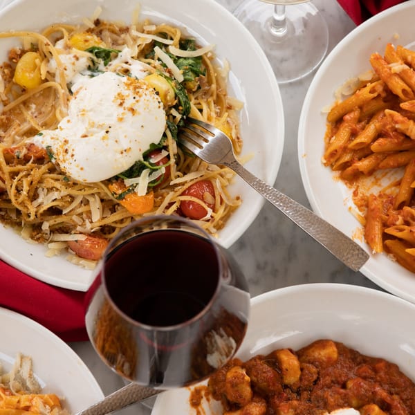 Overhead view of various pasta dishes, including spaghetti with burrata, penne, and gnocchi, with wine glasses.