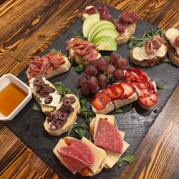 A slate board with various crostini appetizers, including meats, cheeses, fruits, and olives, with a side of honey.