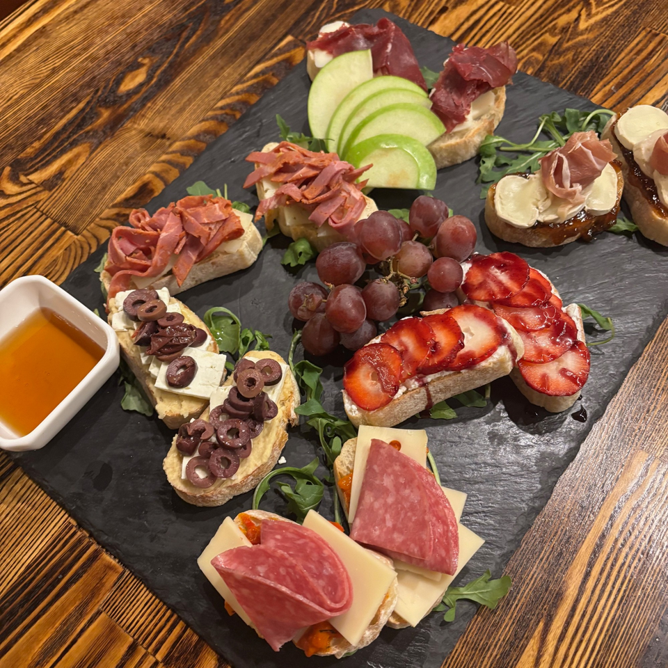 A slate board with various crostini appetizers, including meats, cheeses, fruits, and olives, with a side of honey.