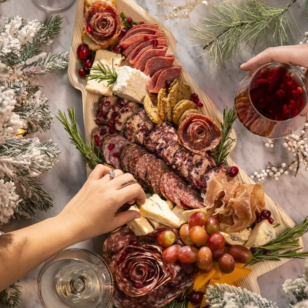 Overhead view of a festive charcuterie board with meats, cheeses, grapes, and rosemary, with hands reaching for food.
