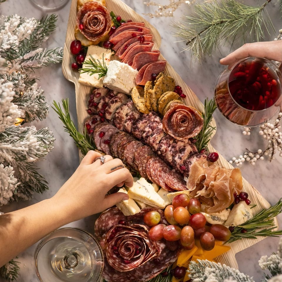 Overhead view of a festive charcuterie board with meats, cheeses, grapes, and rosemary, with hands reaching for food.