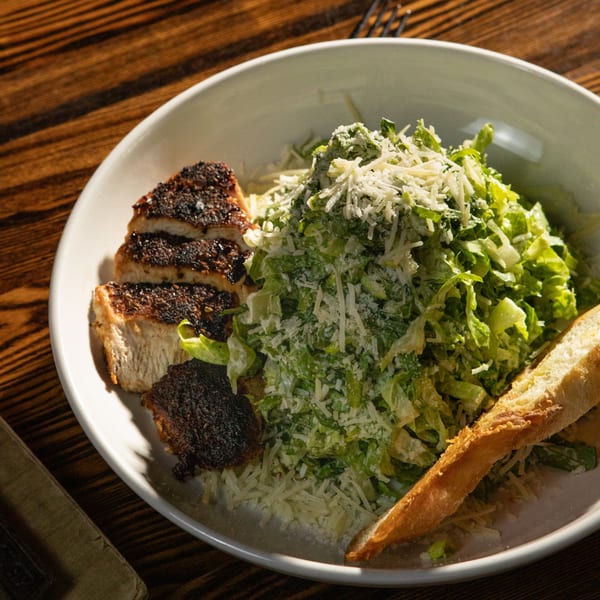 A Caesar salad with grilled chicken and a baguette slice on a white plate.