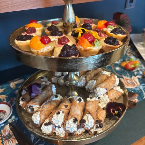 Two-tiered dessert stand with fruit tarts and cannolis, garnished with edible flowers.