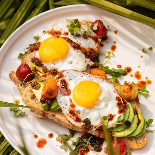 Two avocado toasts with fried eggs, chili oil, cherry tomatoes, and microgreens on a white plate.