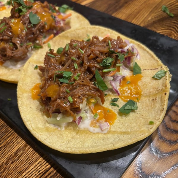 Two Uncommon Grill tacos with shredded meat, slaw, and orange sauce on a black plate.