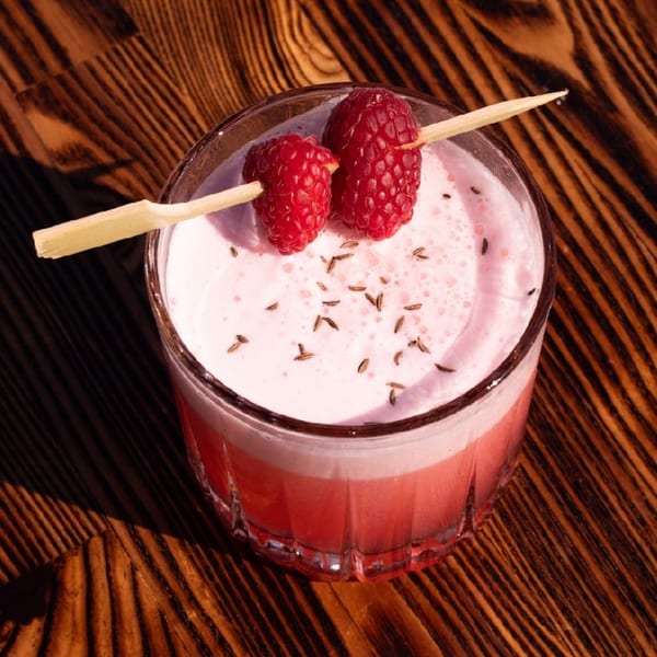Pink cocktail with foam, garnished with two raspberries on a skewer and sprinkled with spices.