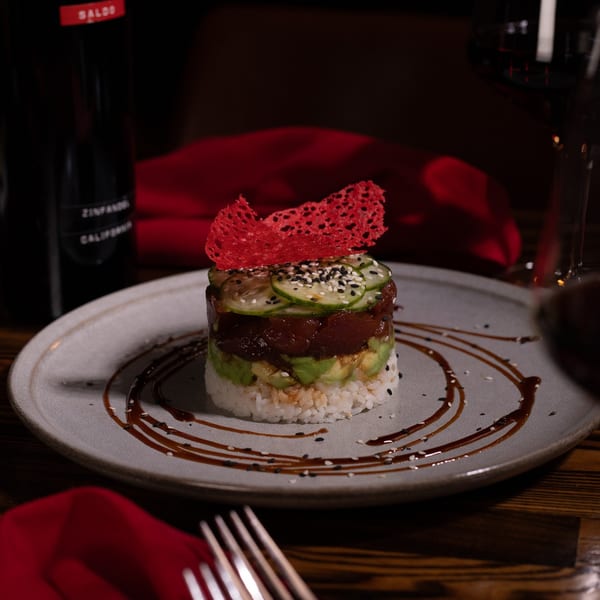 A tuna and avocado tower with cucumber, sesame seeds, and a red crisp, on a plate with sauce.