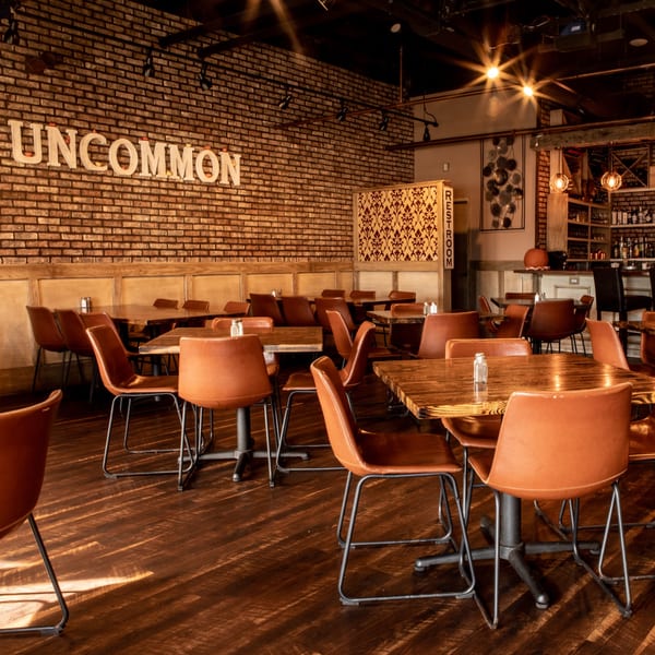 Interior of Uncommon Grill with brick walls, wooden tables, and brown leather chairs, bathed in warm light.