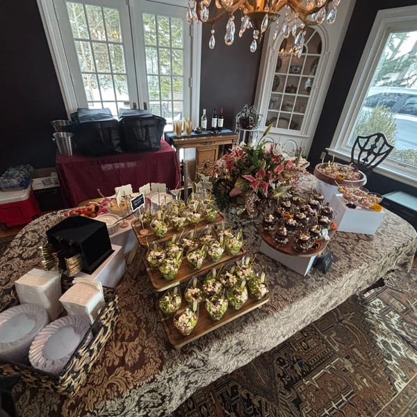 A lavish buffet table with various appetizers, desserts, and drinks, set in a room with large windows.