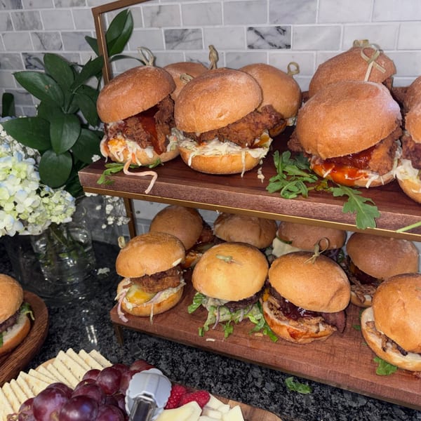 A two-tiered display of Uncommon Grill sliders, with various fillings, alongside a fruit and cheese platter.