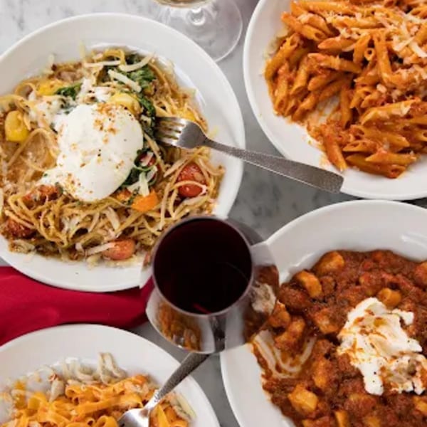 Four bowls of pasta dishes, including spaghetti, penne, and gnocchi, with wine glasses on a table.