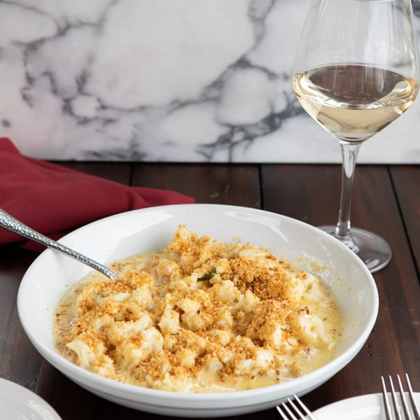 Cauliflower mac and cheese with breadcrumbs in a white bowl, next to a glass of white wine.