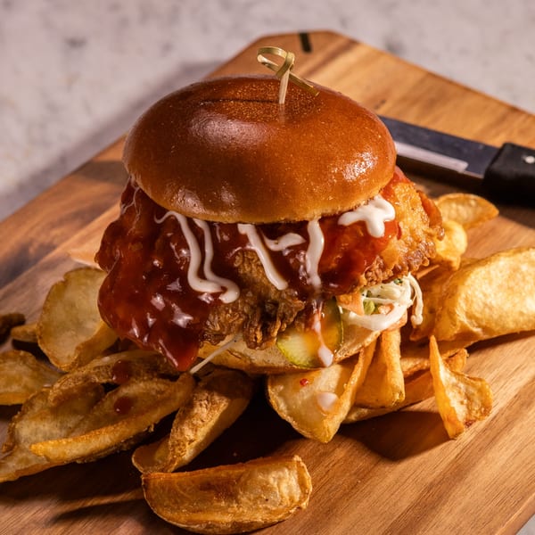 Crispy chicken sandwich with BBQ sauce, coleslaw, and pickles, served with potato wedges on a wooden board.